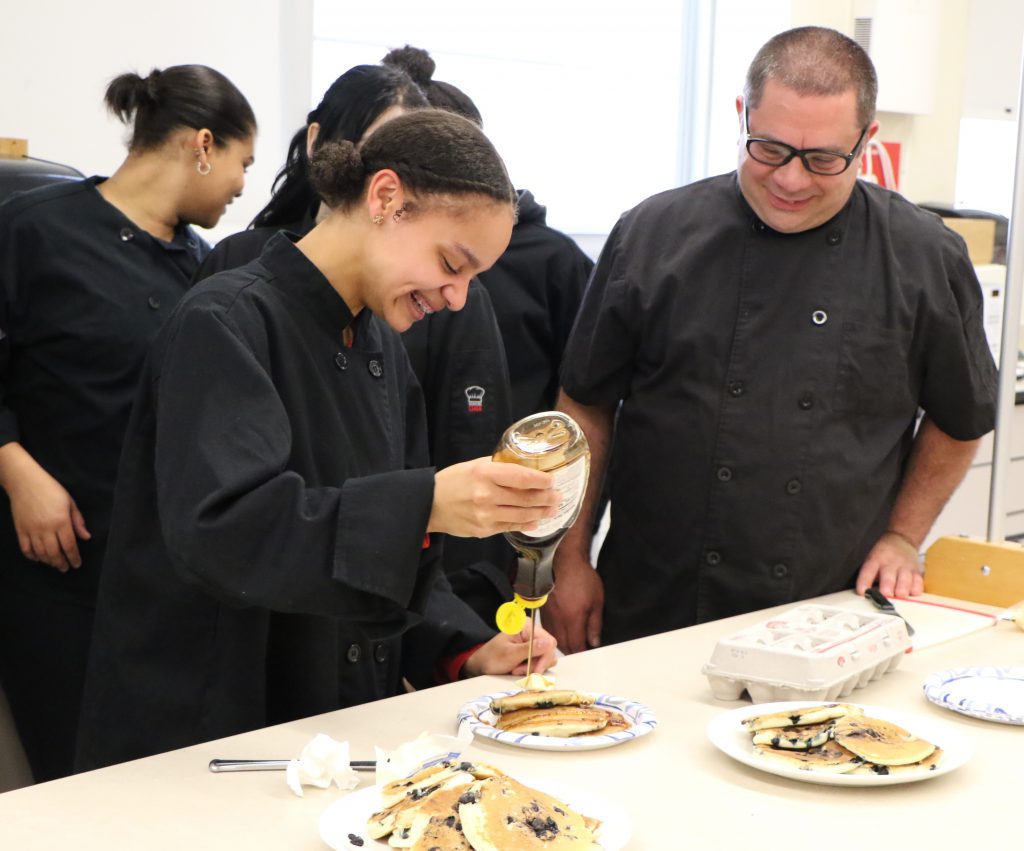 Chef and student in culinary class share a laugh while plating hot blueberry pancakes