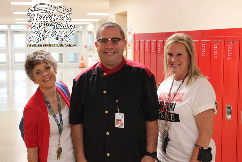 three high school teaching staff members share a smile in the hallway in front of red lockers