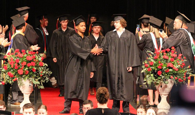 group of graduates in black caps and gowns walking in to the commencement ceremony as teachers and board members clap