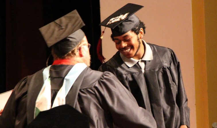 A happy graduate in cap and gown receives his diploma from the school board president