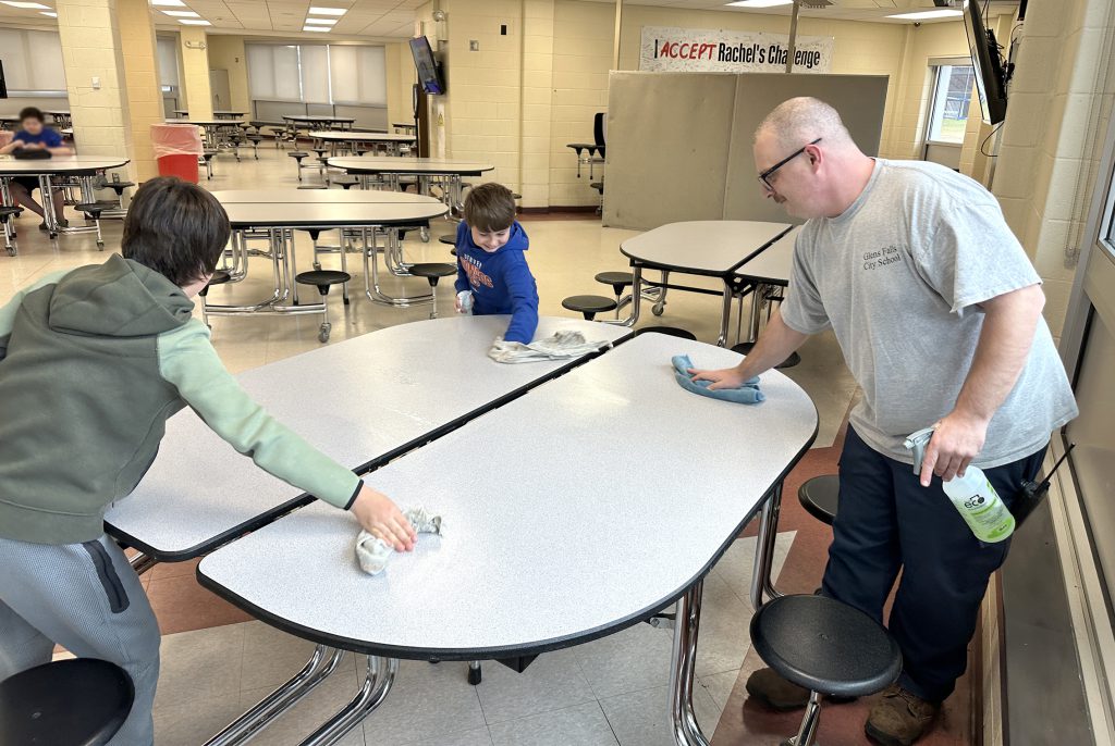 Students helping custodian clean lunch tables with spray bottles and cloths